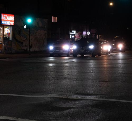 It's dark. On the other side of an intersection, several cars are lined up at the traffic lights; their headlights are brilliant but diffuse, and reflect on the dark pavement, which has distinct ripples in the near field. Behind the cars, miscellaneous white signs. The traffic light is visible, and is green; also there is a white "walk" glyph. In the top right, a street light, also diffuse. In the top center, a curtained window glows. To the left, a H&Z market, a convenience store, glows white …