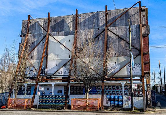 The facade of an under-construction building, propped up with a latticework of rusty steel beams, covered with faded plywood, bare trees in front, complicated shadows, blue sky behind.