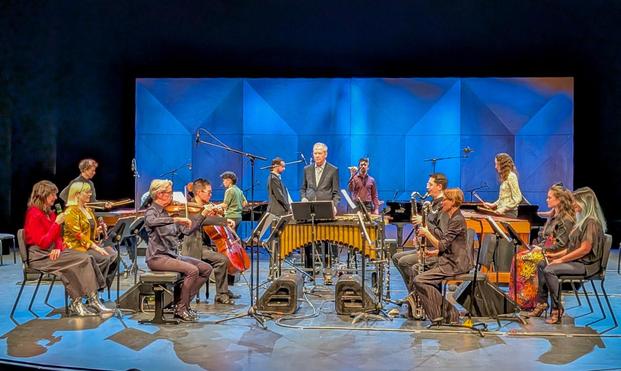 Stage crowded with musicians; at the front we see four instruments being played: violin, cello, and two bass clarinets. Behind those players, seated singers. Many pianos, marimbas, xylophones, and pianos behind them.