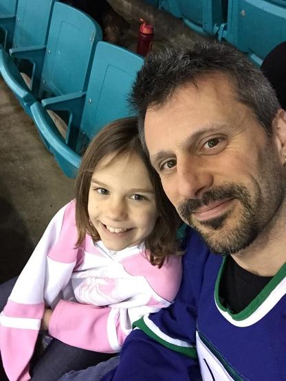 Erin and his youngest daughter, both in Canucks jerseys, his blue, hers pink, smiling at the camera in their seats at a Canucks game.
