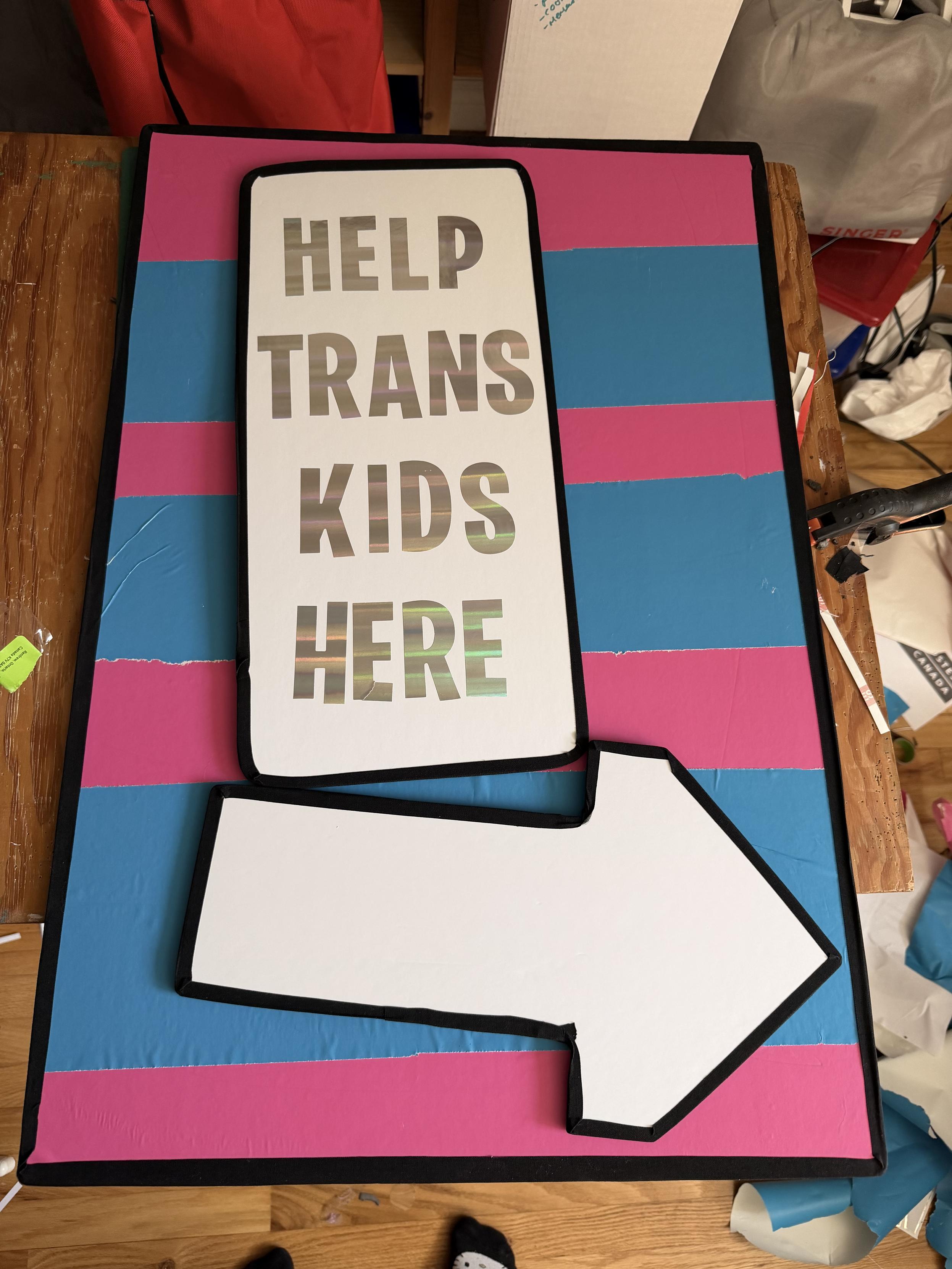 A large sign on pink and blue background that says “help trans kids here” with a large arrow