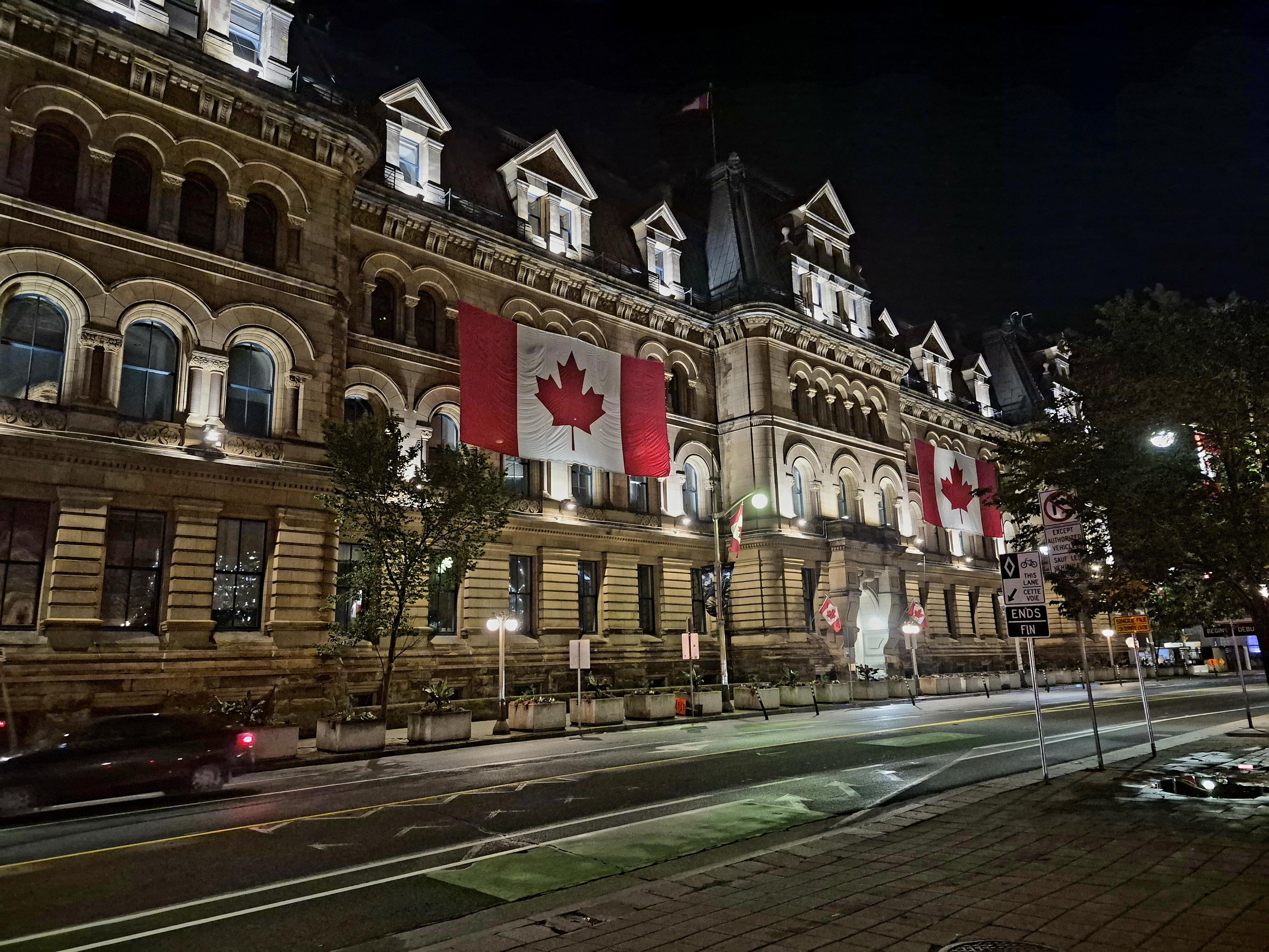 Showing a large roughly 5 story masonry building in a classical style illuminated from the outside at night. There are two large Canadian flags on the outside about mid way up.