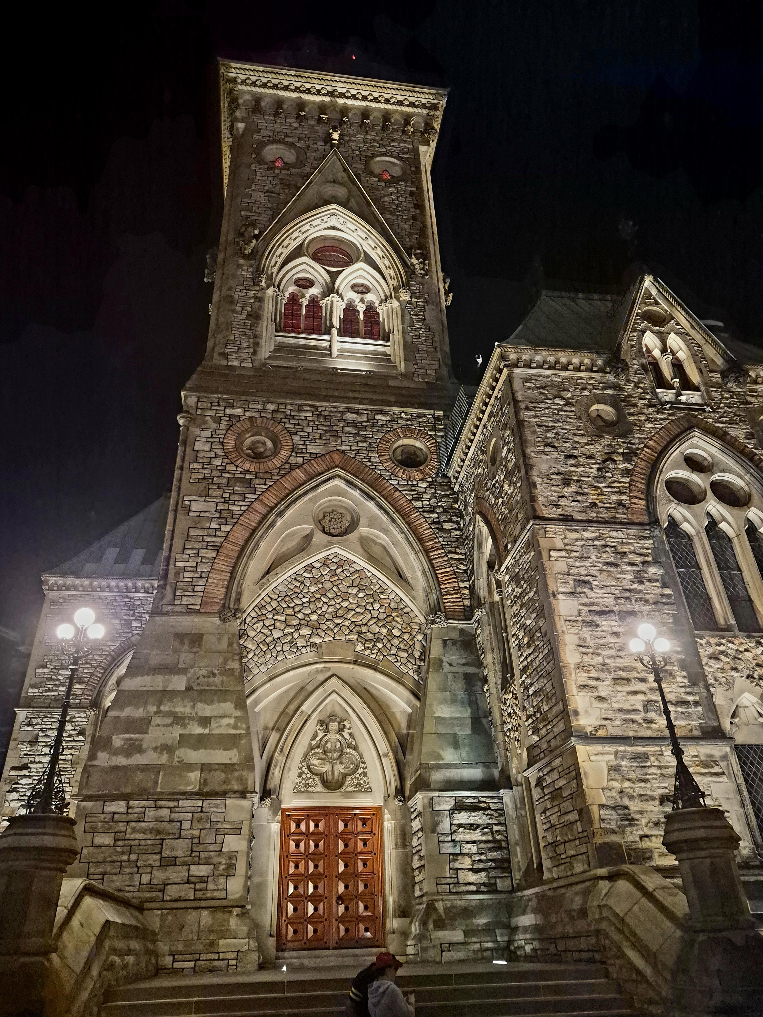 Showing a close up picture of East Block, showing the large entrance and lots of masonry illuminated at night, gothic arches and windows as well as a bell tower.