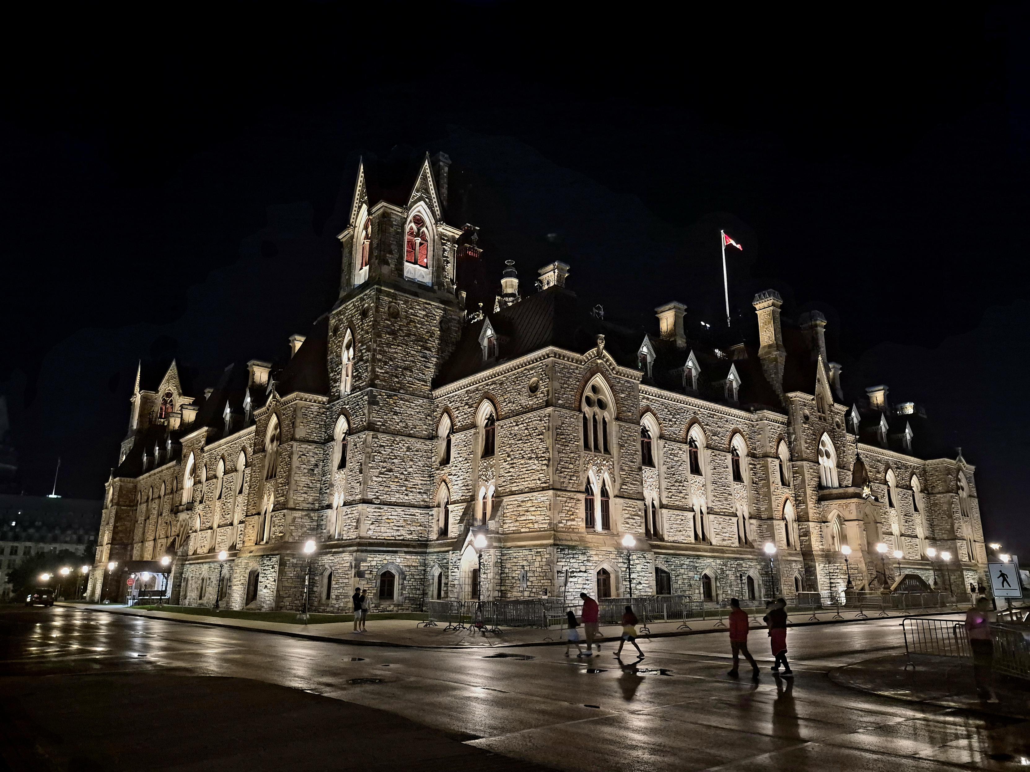Showing the center side of West block, lots of masonry and gothic styled details. There are towers on the corners and mid way, a Canadian flag on a pole is visible, as well as people walking out front.