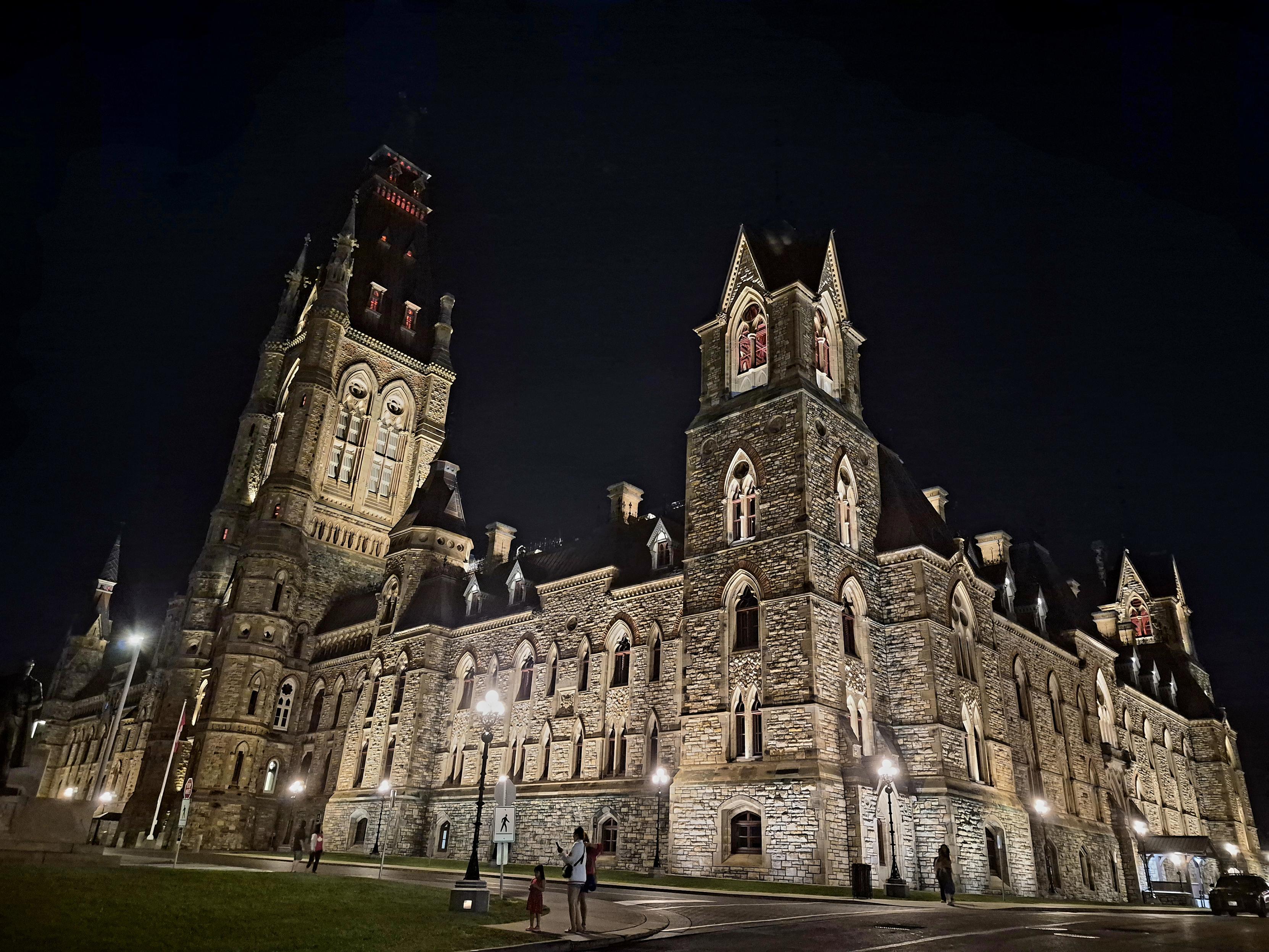 Showing the western corner of West Block, there is more of th same masonry and lights illuminating the gothic style building at night, with a large tower middle of the west side with a large copper roof.
