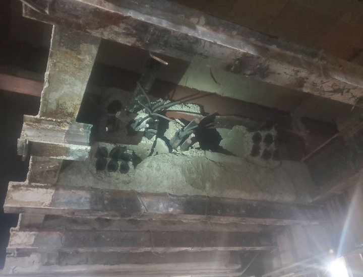 Looking up into a grid of I-beams. Tubes can be seen going into concrete and are all broken off. Ripped cables can be seen sticking out.