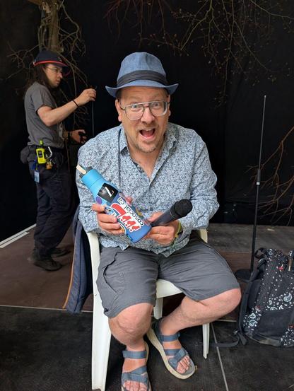 Boris Mann with summer blue hat and blue shirt and shorts. Sitting in a chair holding a water bottle with many stickers - including cosocial.ca