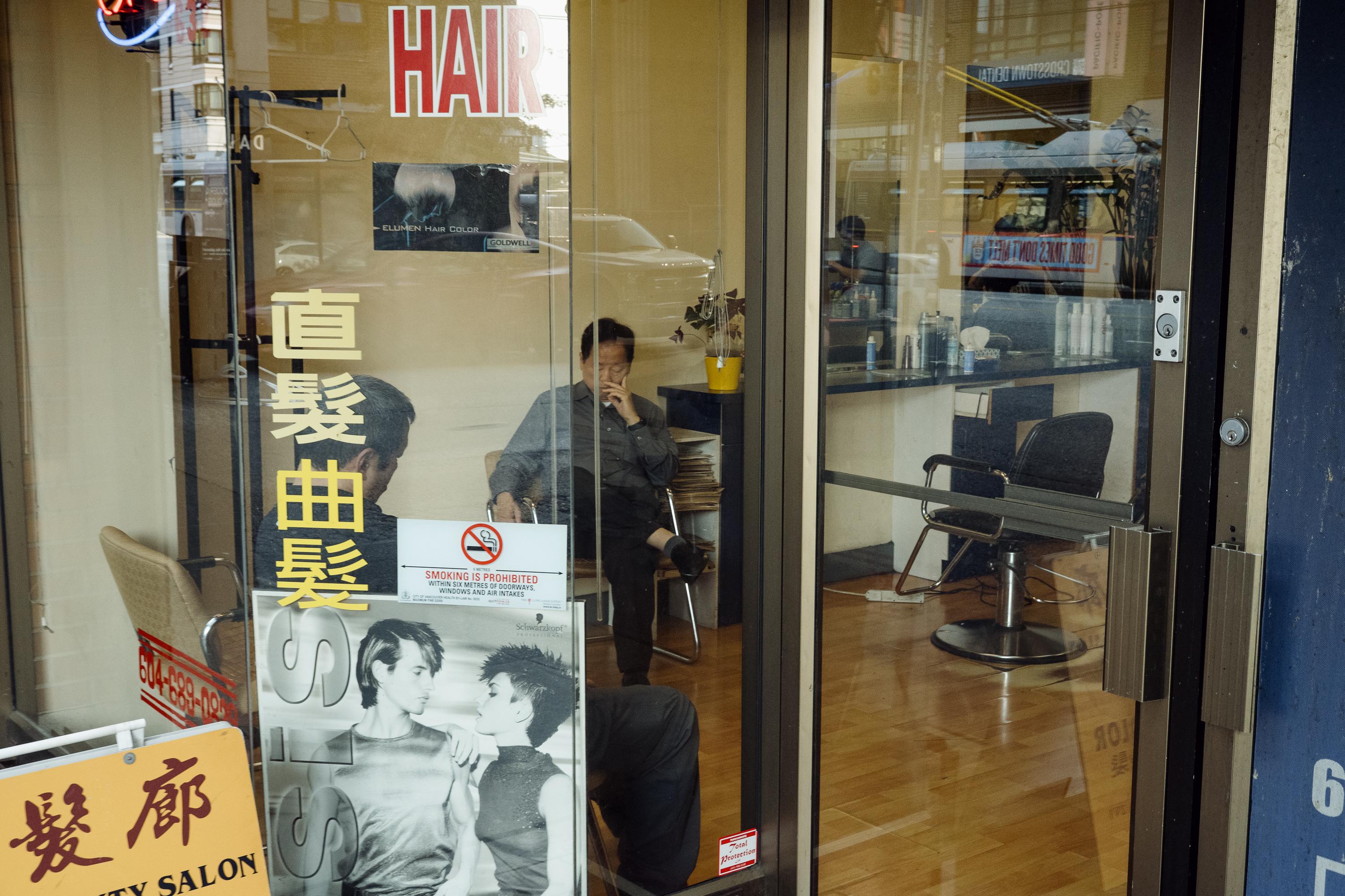 The interior of a Chinatown hair salon viewed through its glass storefront. Chinese characters and the word HAIR are painted on the window. Two people are visible inside — one seated in a stylist's chair, another resting in a waiting chair. A Schwarzkopf hair colour poster and a no smoking sign are affixed to the glass.