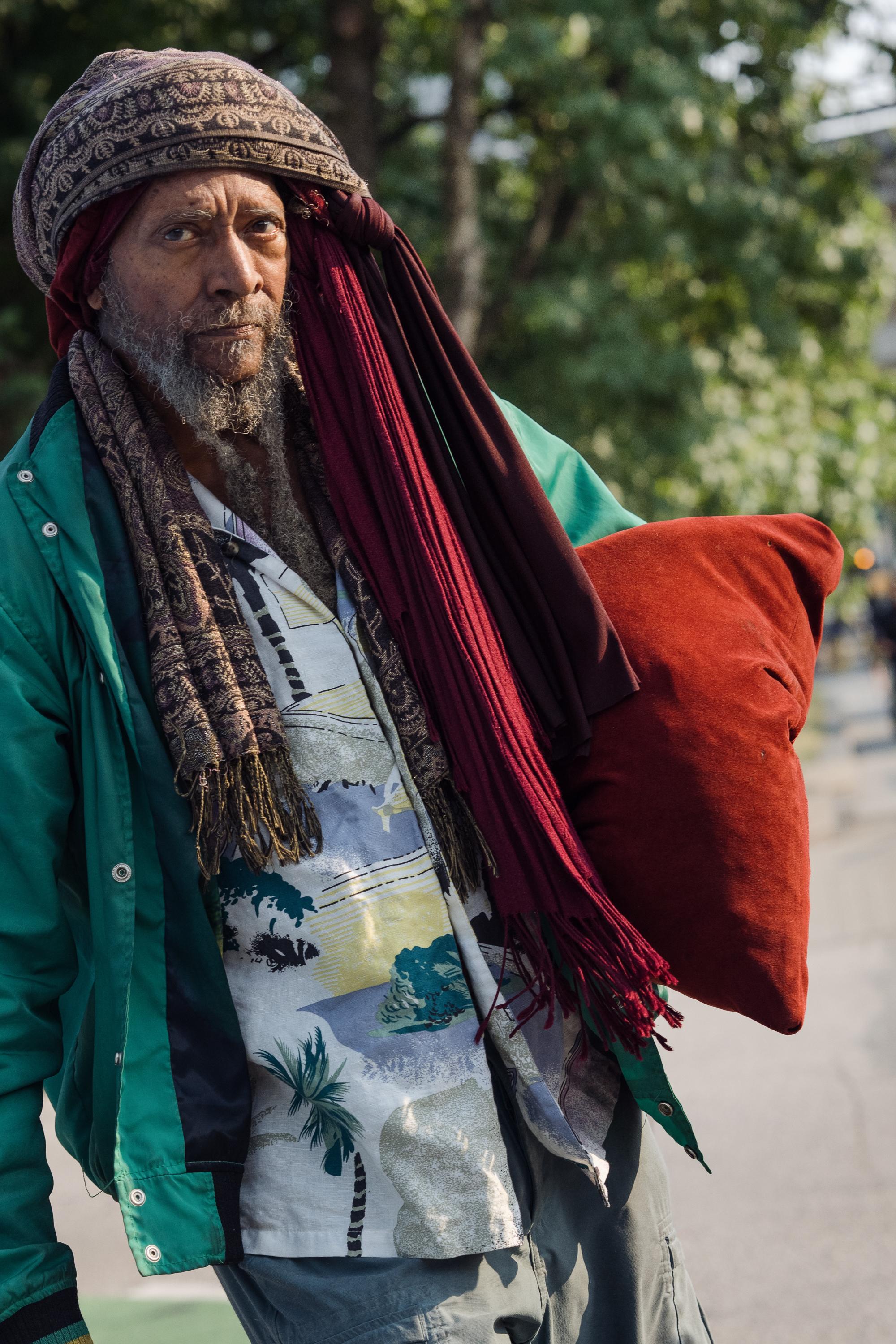 An older man stands on a Vancouver street looking directly at the camera with a calm, steady gaze. He wears a patterned head wrap, a long burgundy scarf, a floral shirt, and a green jacket, and carries a large rust-coloured cushion under one arm. Trees are softly blurred behind him.