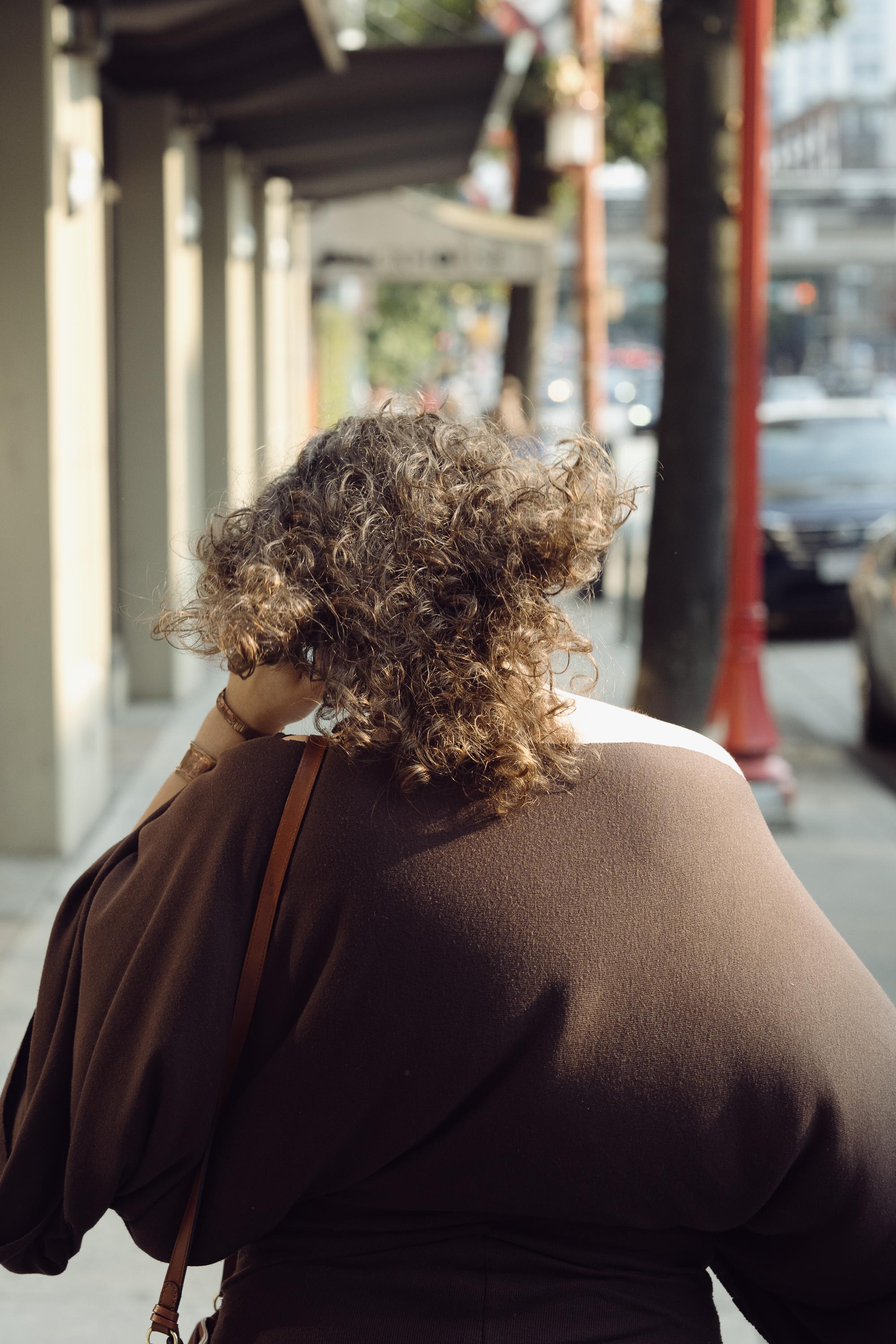 A woman walks away from the camera along a covered sidewalk in Vancouver's Chinatown. Her curly hair catches the warm light. A red ornamental lamp post and parked cars are visible in the background.
