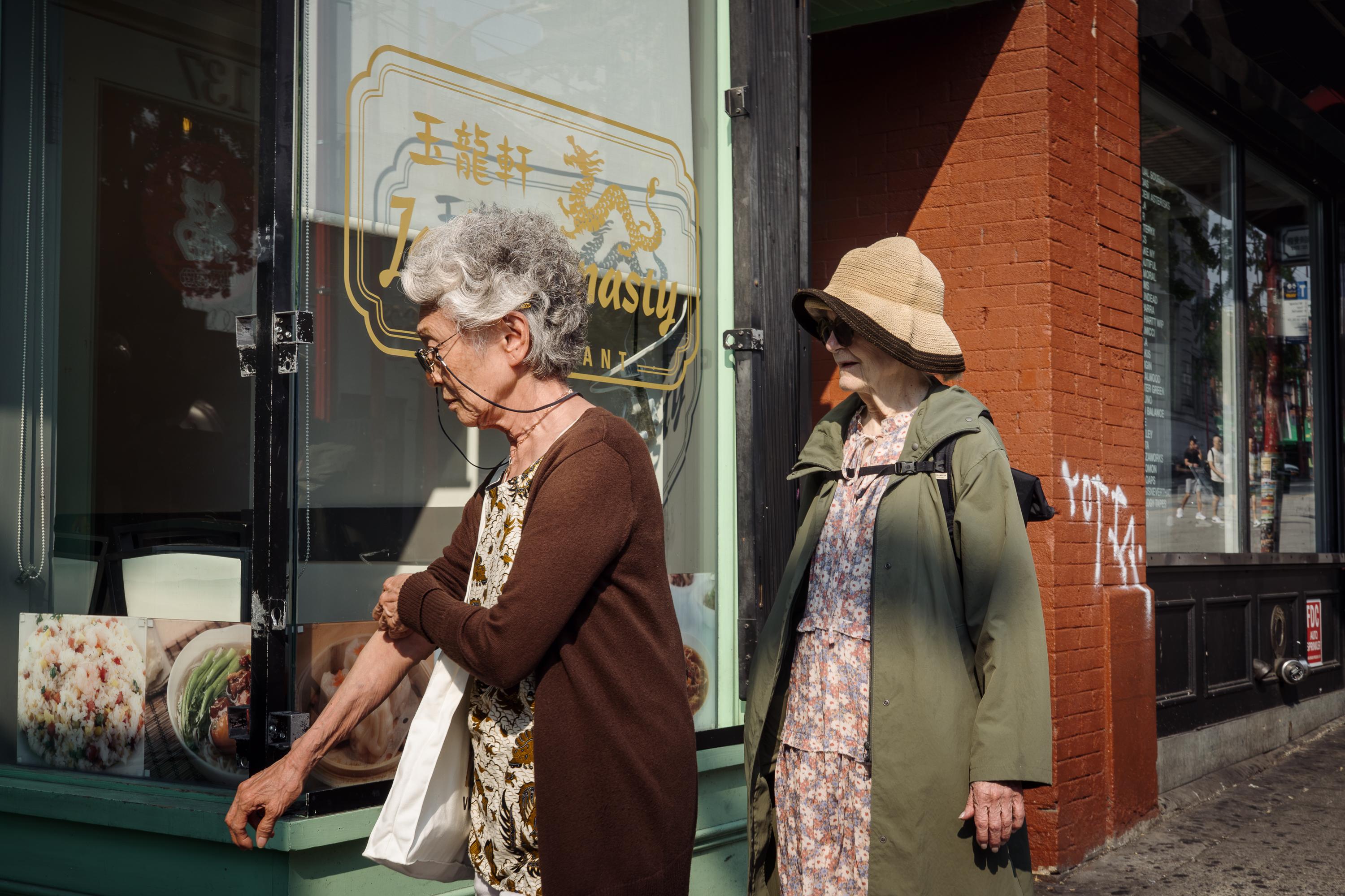 Two older women stand on the sidewalk outside a Chinese restaurant in Vancouver's Chinatown. One, with short grey hair and glasses, leans toward the restaurant window carrying a white tote bag. The other, wearing a wide-brimmed straw hat, green coat, and floral dress, stands beside her looking ahead. Warm sunlight falls across the red brick wall behind them.