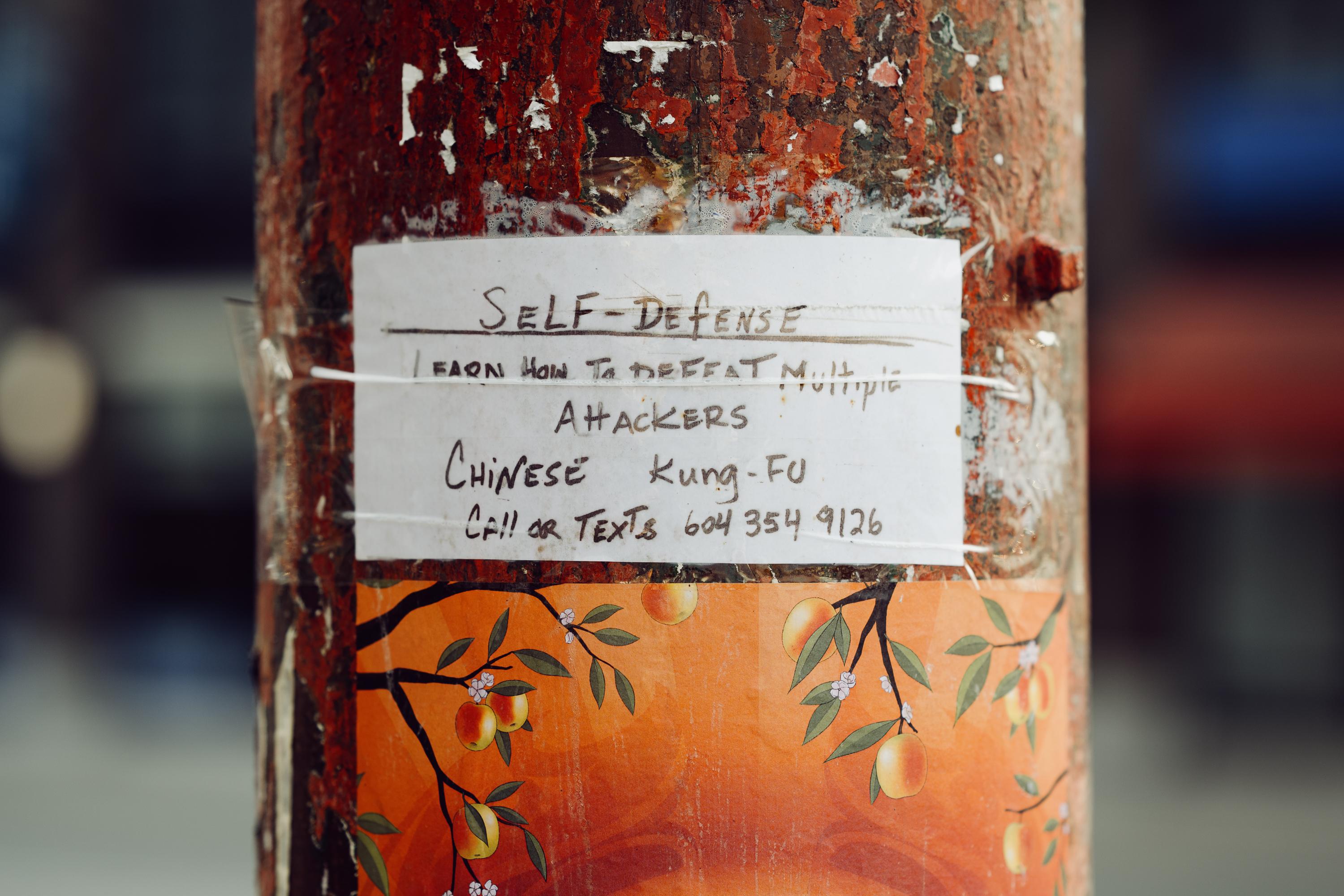 A handwritten flyer taped to a weathered, paint-peeling pole in Vancouver's Chinatown advertises self-defense classes in Chinese kung fu, promising to teach how to defeat multiple attackers. Below the flyer, the pole is painted with an orange mural of fruit-laden branches.