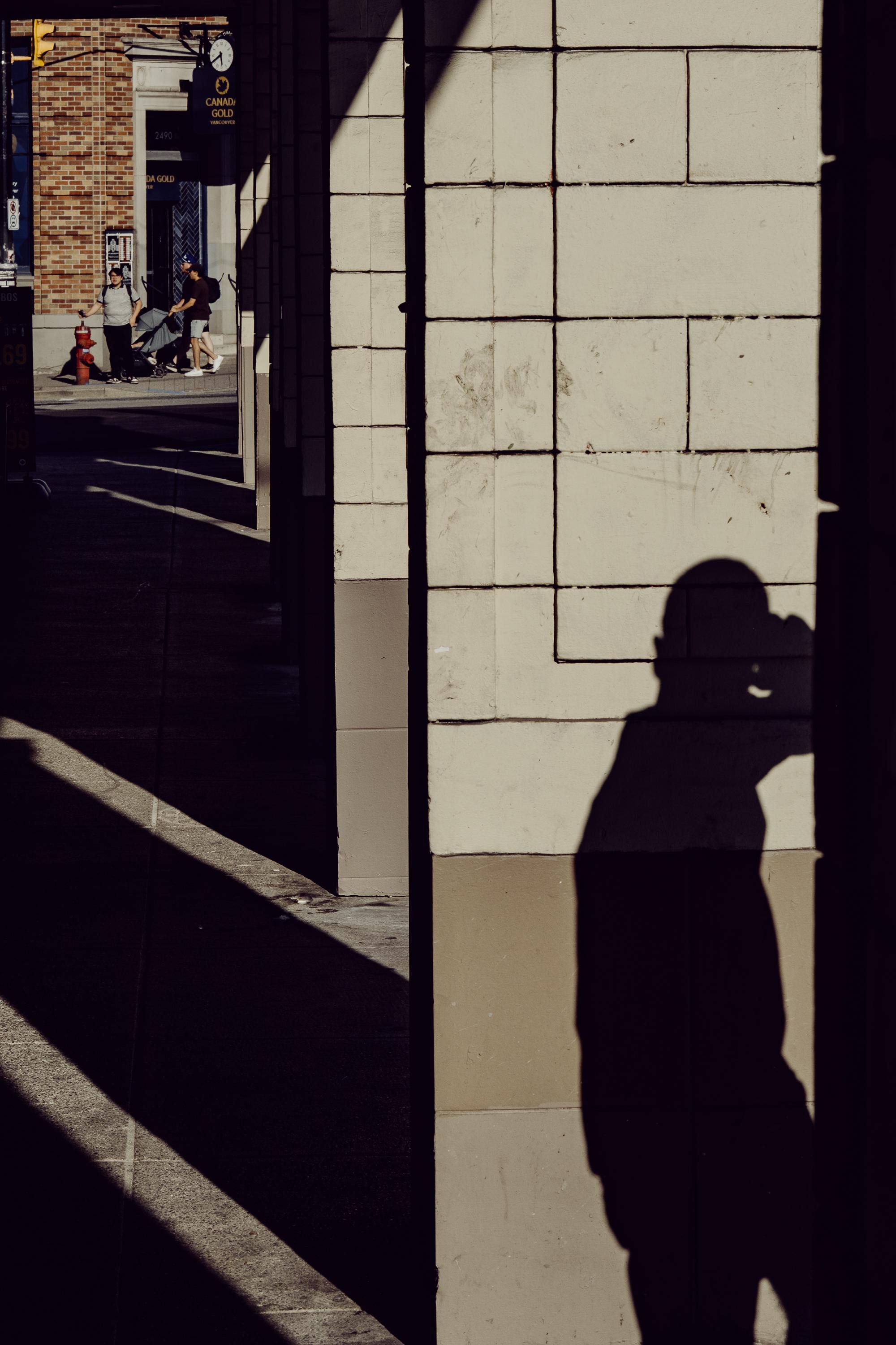 A deep shadow falls across a tiled building facade on a Vancouver sidewalk, casting the sharp silhouette of a person raising a camera to their eye. Through a gap between buildings in the background, a small group of people stand on a sunlit street beside a fire hydrant. A Canada Gold sign and a clock are visible in the distance.