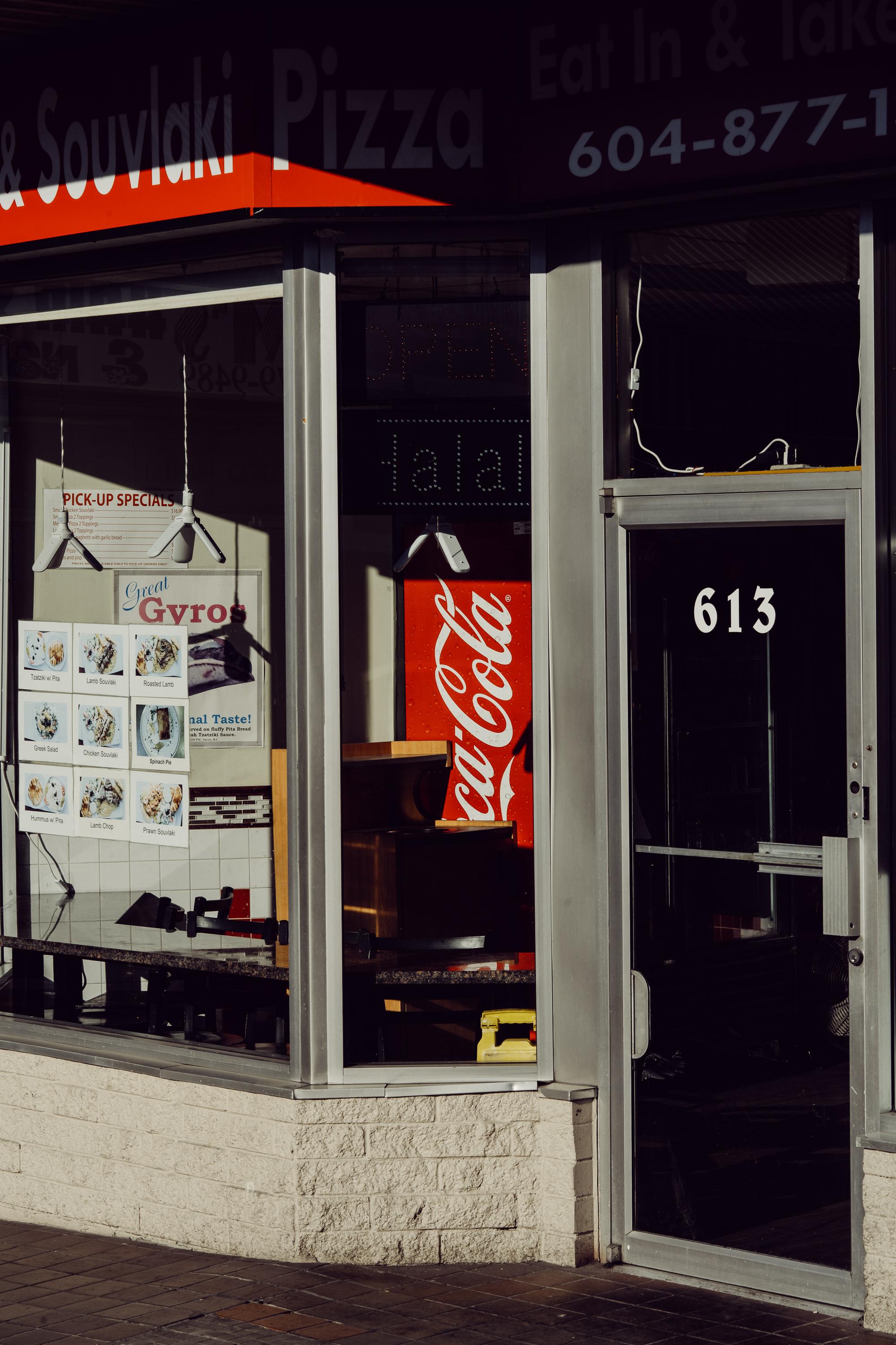 The storefront of a souvlaki and pizza restaurant on a Vancouver street, lit by strong directional sunlight. A Coca-Cola fridge and a Great Gyros menu poster are visible through the window. The number 613 is displayed on the door.