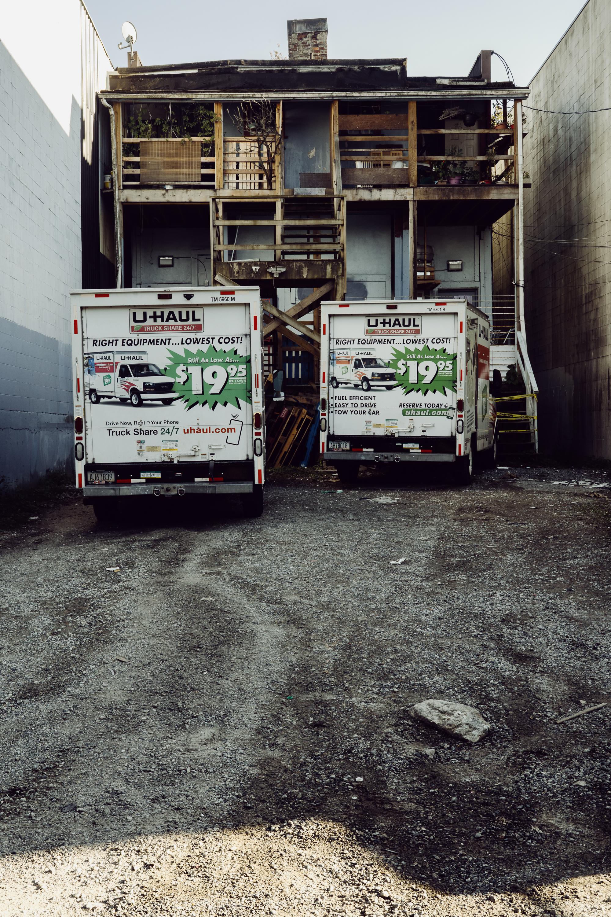 Two U-Haul trucks parked side by side in a gravel back alley, their identical rear advertising panels facing the camera. Behind them, a weathered two-storey wooden building with open staircases and balconies rises between two concrete walls.