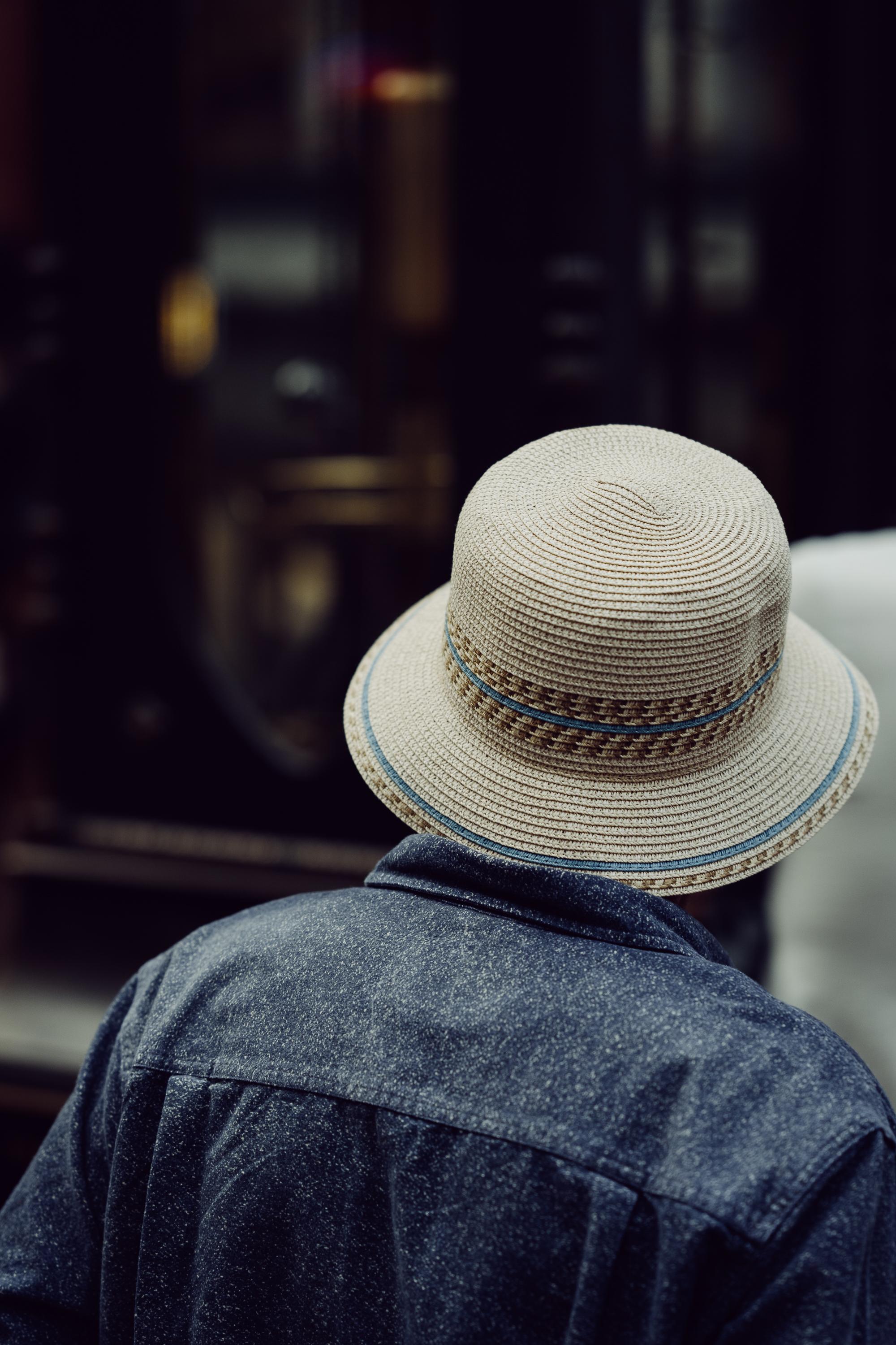 Close-up of the back of a person's head on a Vancouver street. They wear a woven straw bucket hat with a blue band and a dark denim jacket. The background is dark and softly blurred.