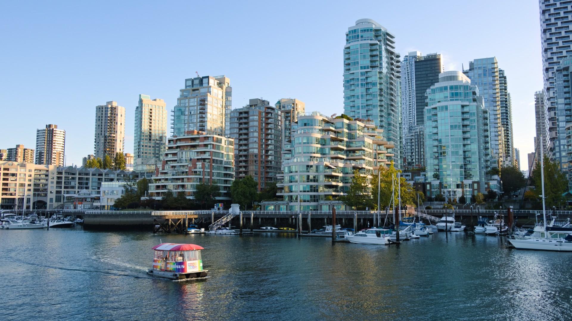 Scene of a harbour, with downtown Vancouver in the near distance. A small ferry is crossing from left to right. This ferry has a rainbow design on the sides and a Canada flag design on top. Morning light, clear skies.