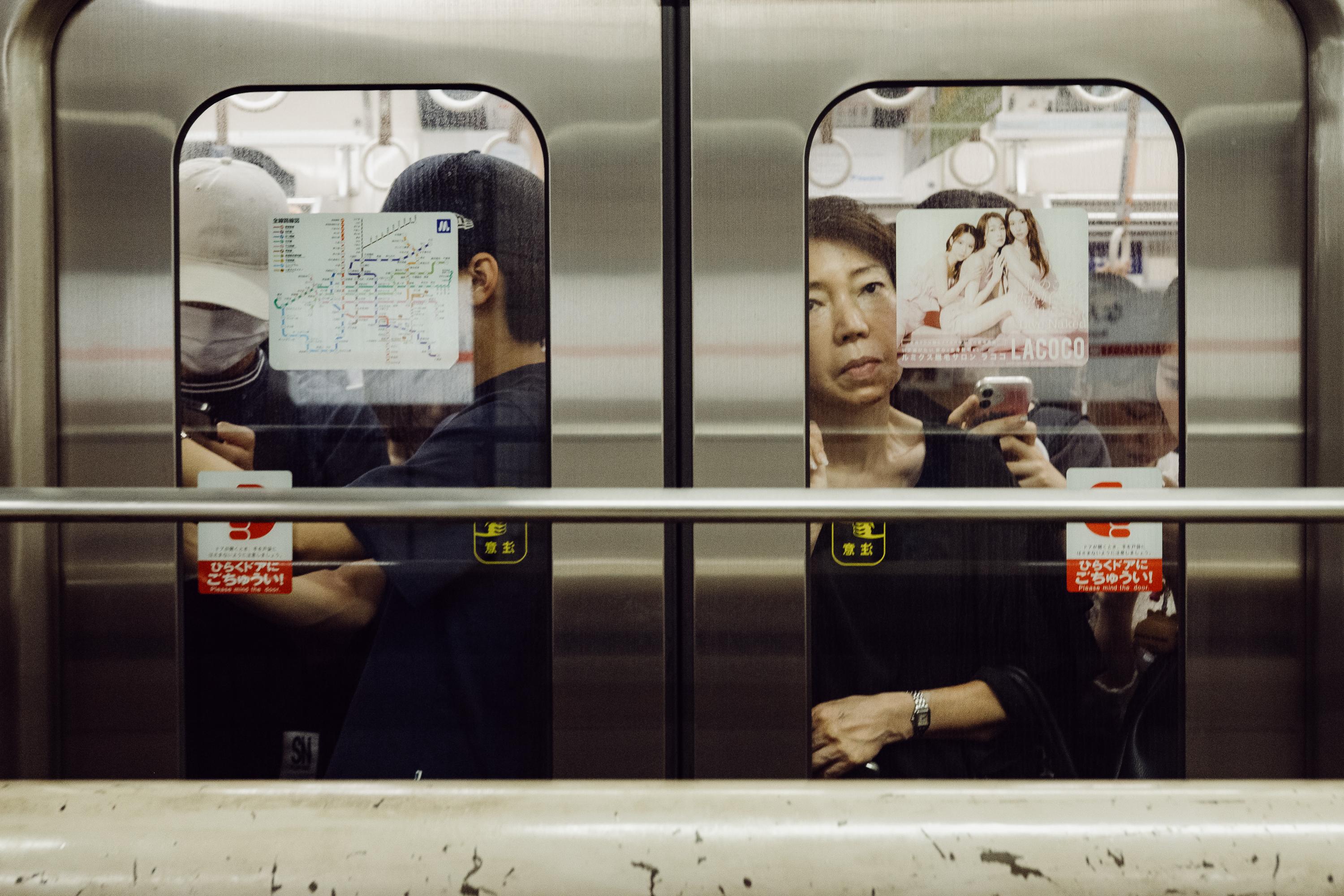 A woman seated in a Tokyo subway car looks directly at the camera while holding her phone. Beside her, the back of another passenger's head is visible. A metro map and an advertisement are posted on the glass doors between them.