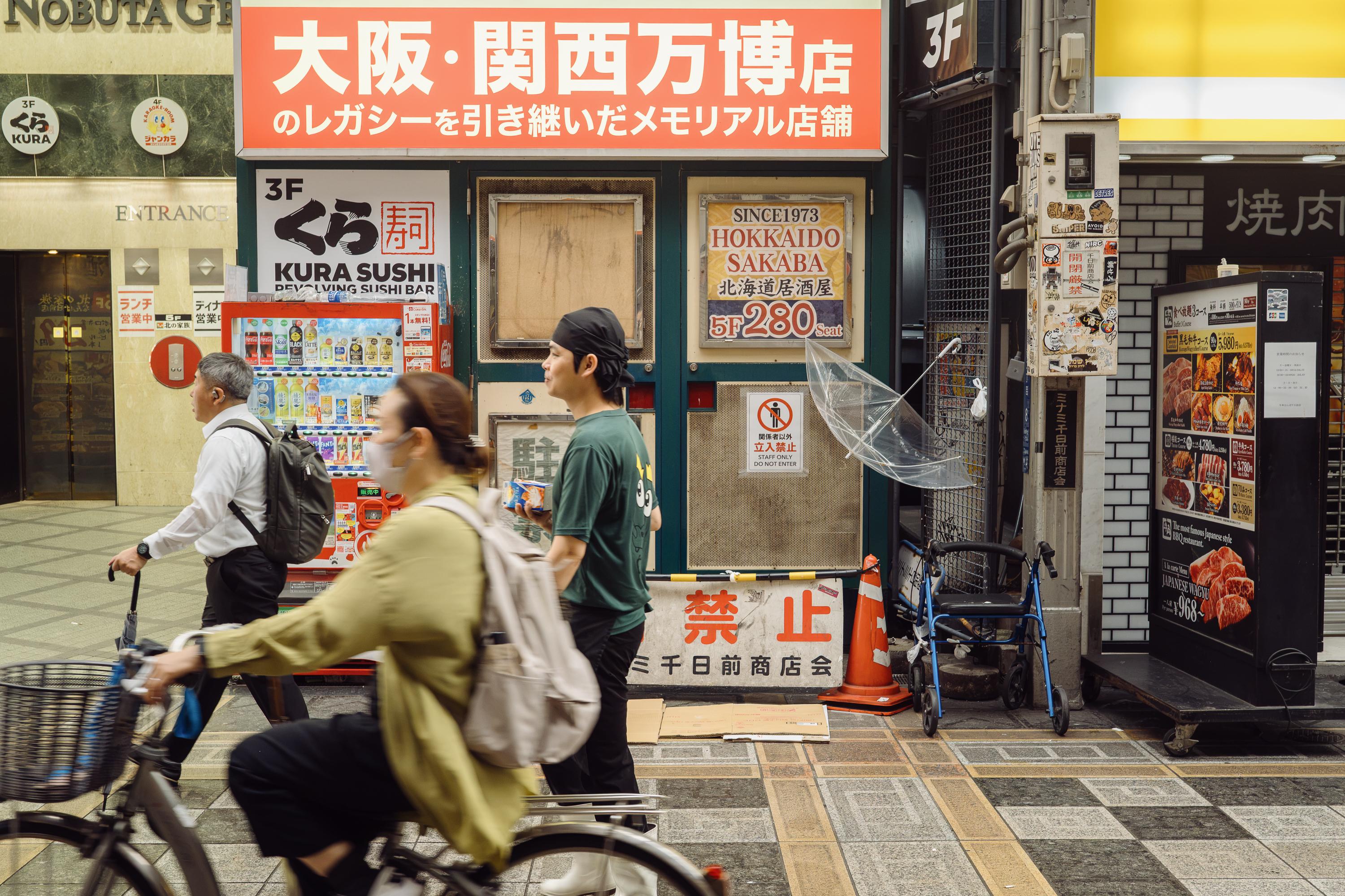 A busy covered shopping arcade in Osaka. A woman cycles past in the foreground while a man in a white shirt walks behind her. A large orange sign with Japanese characters advertises the Osaka Kansai Expo memorial store. A Kura Sushi revolving sushi bar sign is visible, along with a vending machine and scattered street furniture.