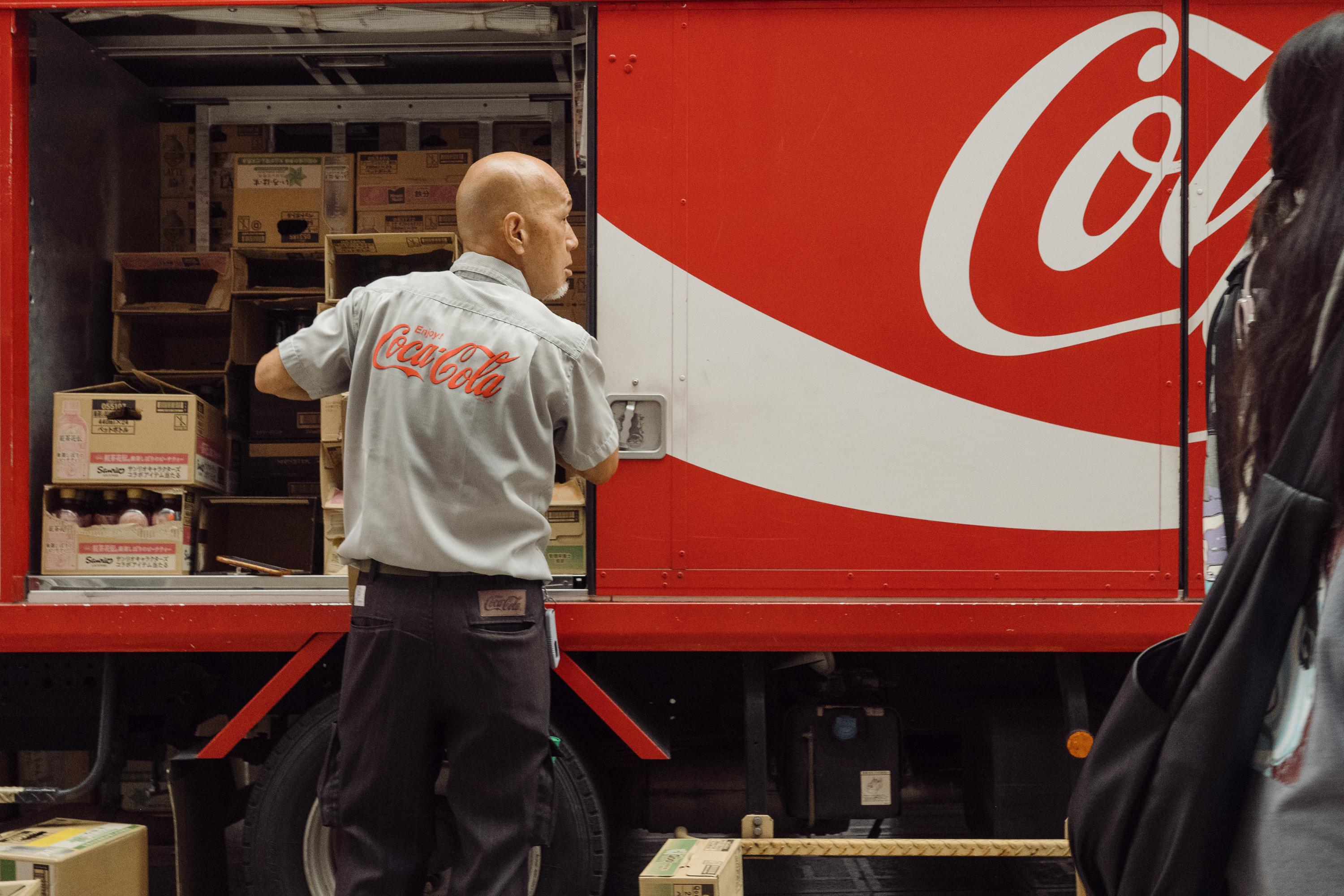 A Coca-Cola delivery worker in a branded grey uniform stands at the open rear doors of a large red Coca-Cola delivery truck, unloading cardboard boxes. The Coca-Cola logo in red and white fills the side of the truck.