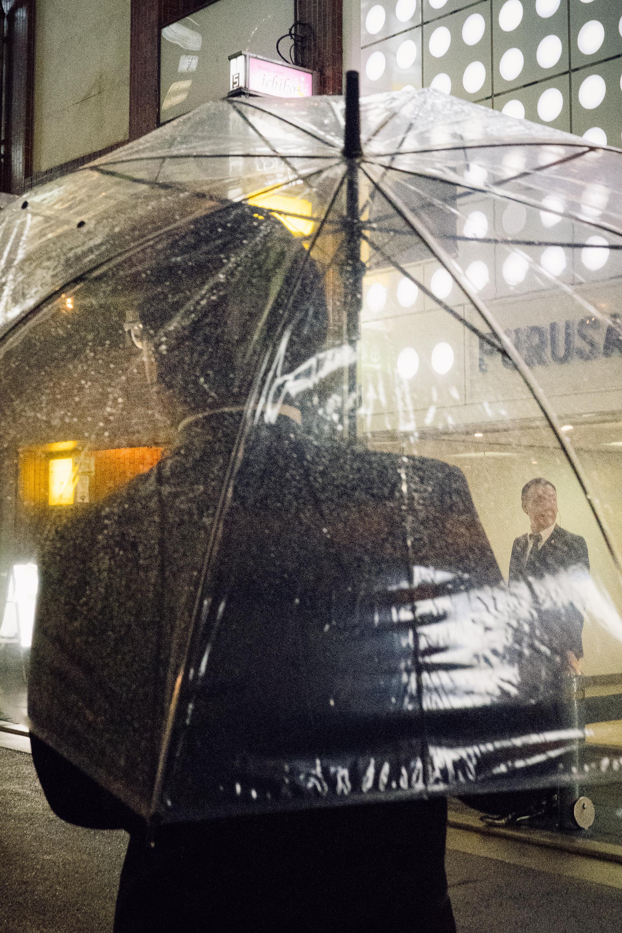 A person holding a large transparent dome umbrella stands on a rain-soaked street in Osaka at night. Through the umbrella, the lights of surrounding buildings are visible, along with a smiling man in a suit.
