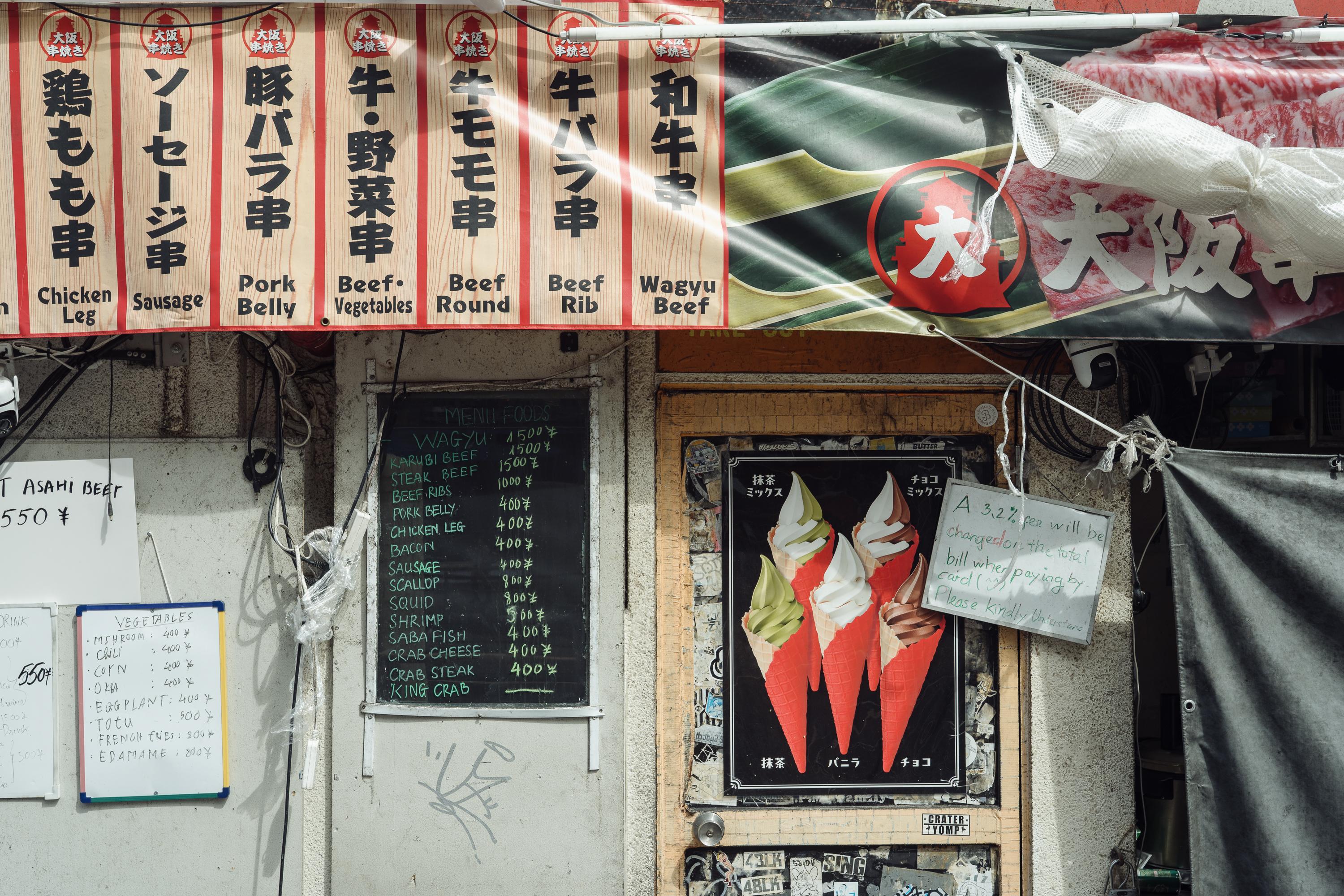A cluttered Osaka kushikatsu restaurant facade. A banner across the top lists skewer menu items in Japanese and English including chicken leg, sausage, pork belly, beef and wagyu. Handwritten chalk menus cover the walls, alongside a poster showing soft-serve ice cream cones.