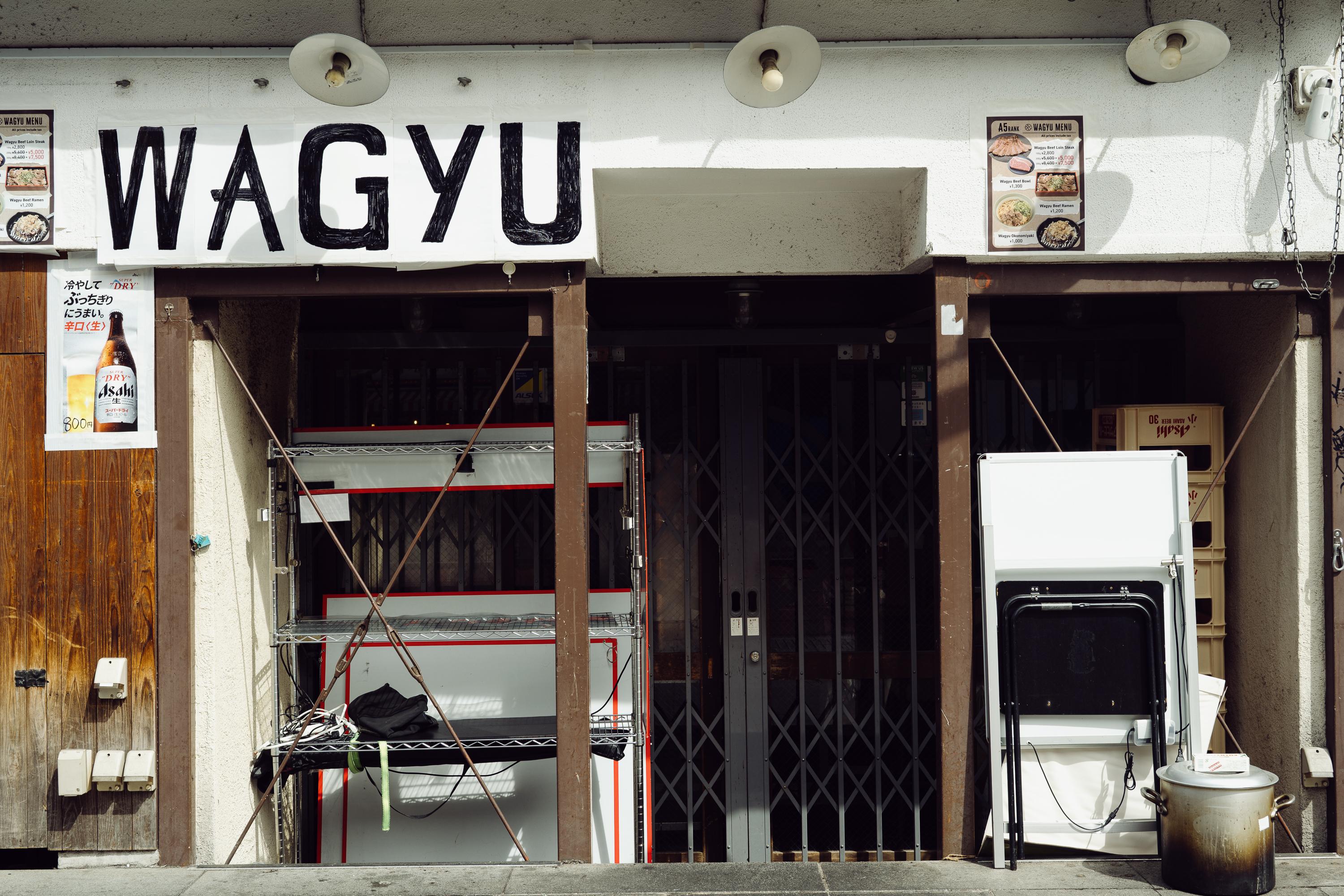 The closed facade of a Wagyu restaurant in Osaka. A large hand-painted black sign reading WAGYU spans the width of the building. Wire shelving and miscellaneous items are visible through the open shutters. Menu boards advertise wagyu beef dishes and Asahi beer.
