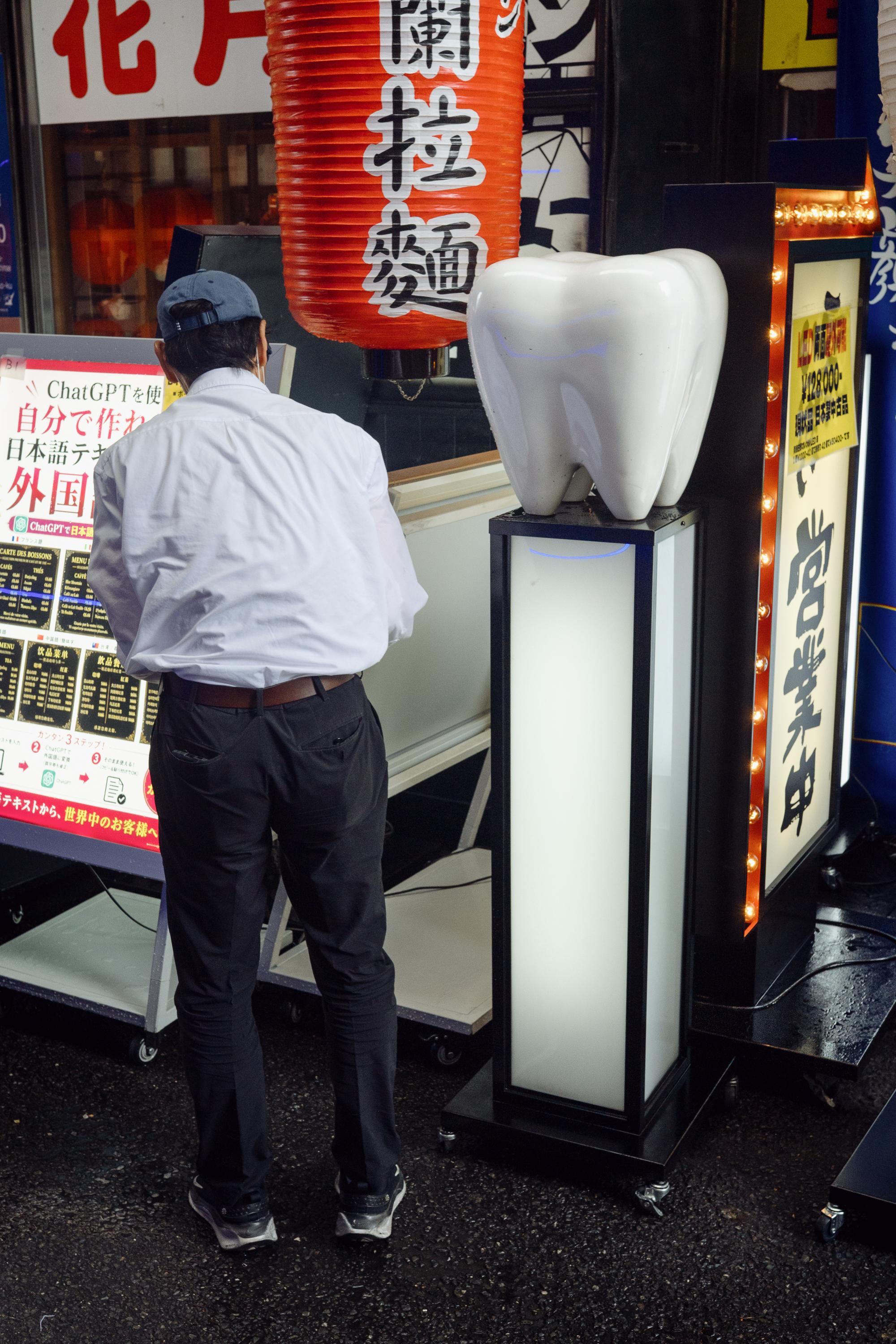 A man in a white shirt and dark trousers stands with his back to the camera on a busy Osaka street, reading a sign. Beside him, a large white sculpture of a tooth sits atop a illuminated pedestal. A red paper lantern and Japanese signage are visible in the background.