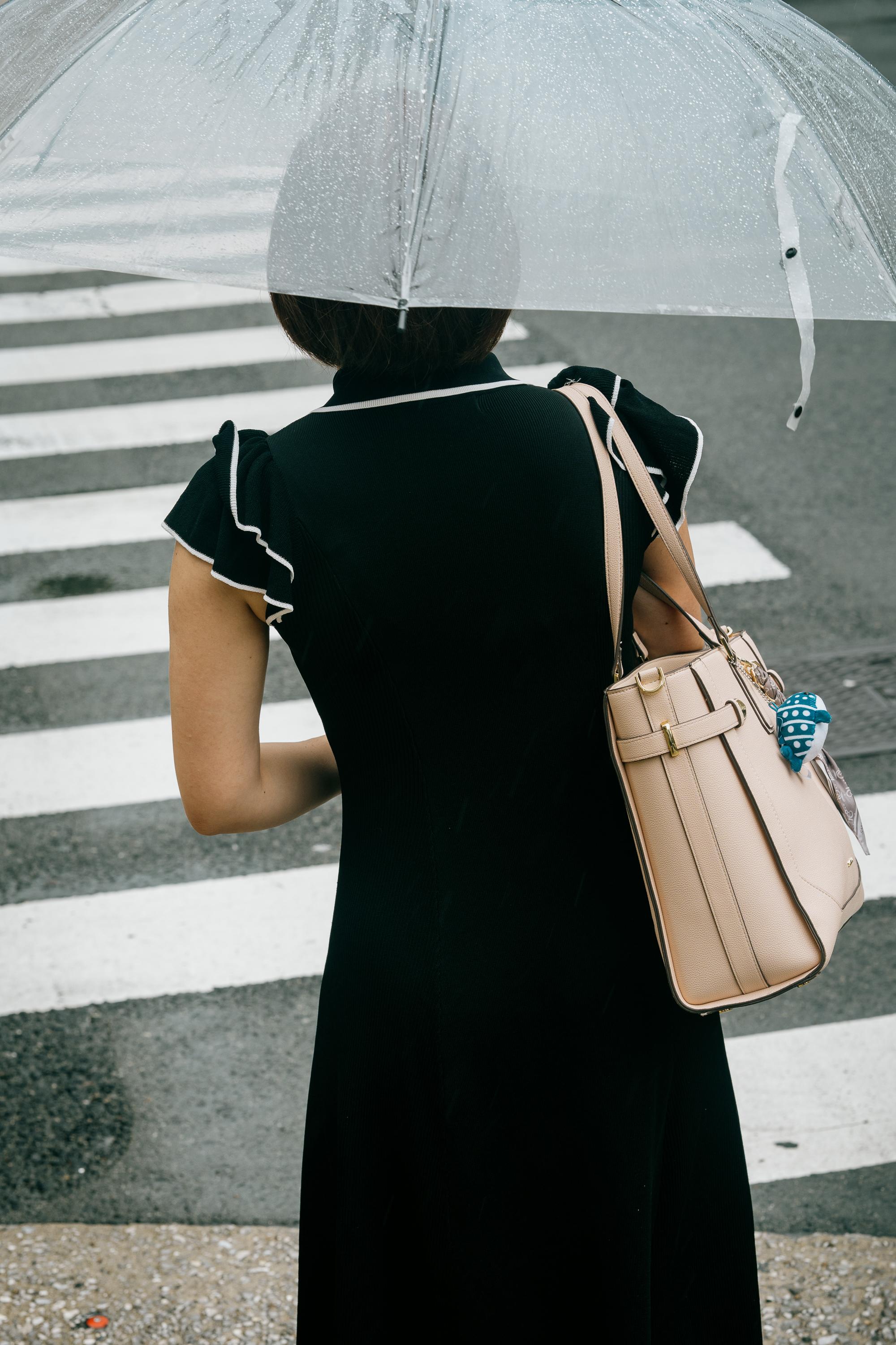 A woman stands with her back to the camera at a pedestrian crossing in Osaka in the rain. She holds a clear dome umbrella and carries a beige leather handbag. She wears a black dress with ruffled cap sleeves edged in white. Bold white zebra crossing stripes fill the background.