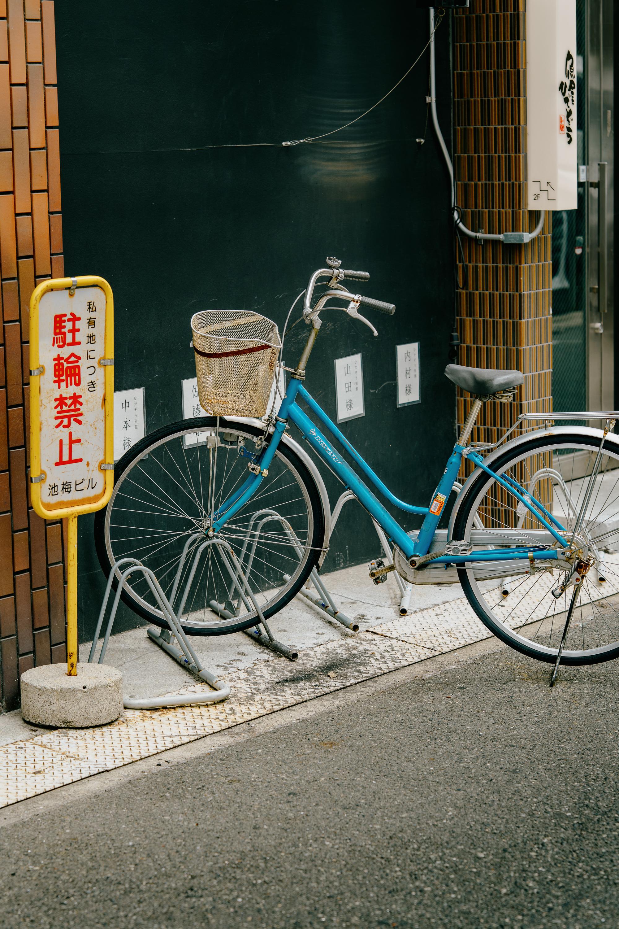 A blue city bicycle with a front basket is parked against a dark tiled wall on an Osaka sidewalk, next to a yellow and white no-parking sign in Japanese. Name plaques are mounted on the wall behind it.