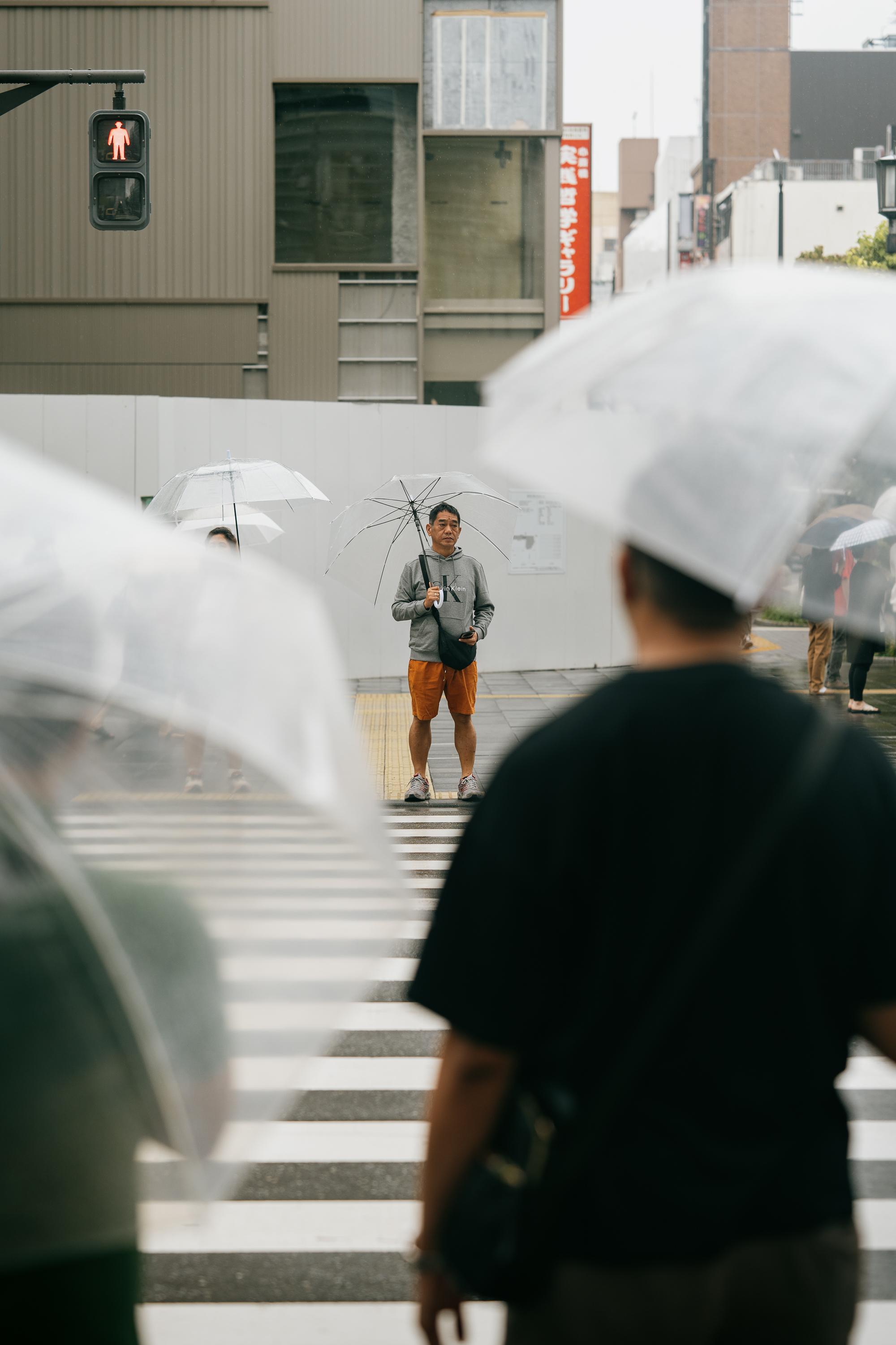 Pedestrians wait at a crosswalk in Osaka on a rainy day, most holding clear dome umbrellas. A man in a grey hoodie and orange shorts stands in the centre of the frame, facing the camera. A red pedestrian signal is visible above. Transparent umbrellas fill the foreground and background.