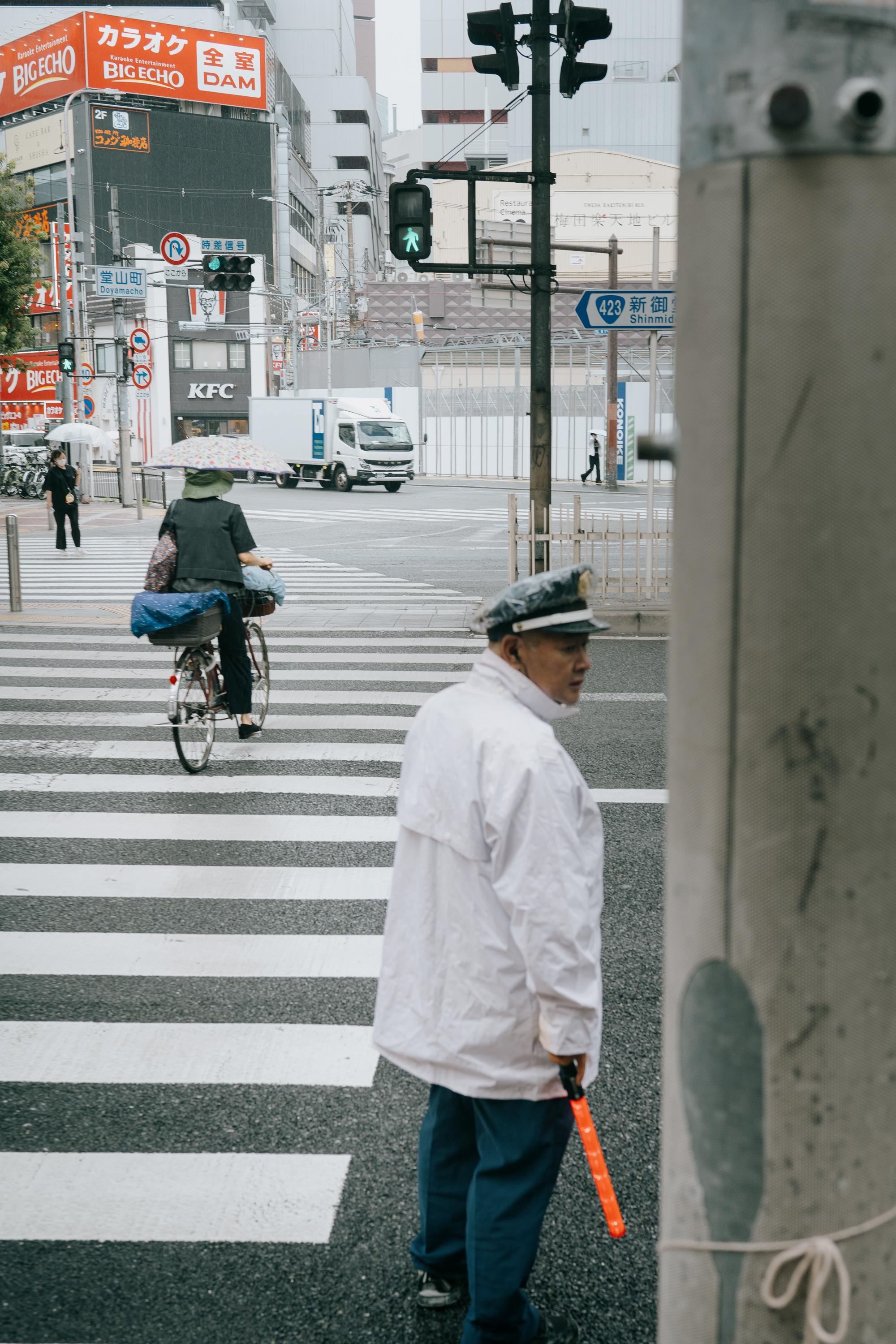 A traffic controller in a white jacket and peaked cap holds an orange baton at a busy Osaka intersection. A cyclist with an umbrella crosses the zebra crossing behind him. Big Echo karaoke signage, a KFC, and a green pedestrian traffic light are visible in the background.