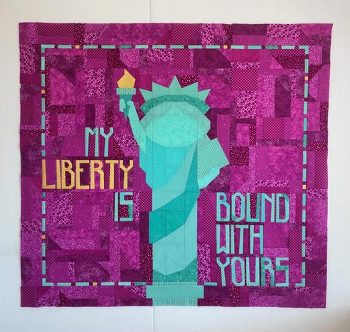 Statue of Liberty-themed quilt, shown in full on a design wall. The somewhat cartoonishly-styled statue is pieced from various coppery green fabrics, though her torch flame is metallic gold. The background is scrappily pieced from a number of tone-on-tone small print fuchsia fabrics. Pieced letters read ‘My liberty is bound with yours” with liberty in the gold metallic and the other words in coppery green. Part of the phrase is on her right side and part is on her left and the sections are linked by a dashed coppery green border that encircles the words and statue on all four sides. A medium wide scrappy fuchsia border finishes off the quilt.