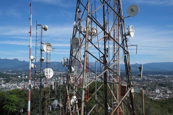 Photo of part of a large telecoms tower, taken from a high elevation, as a city is seen below spread across the landscape with mountains behind. The tower consists of several small and large masts, upon which numerous satellite discs are mounted. The cityscape far below is nestled in a landscape of green forest in the foreground and blue mountains behind. The sky is blue and clear. Joinville, Brazil, 3 October 2017. Photo Cindy Kohtala