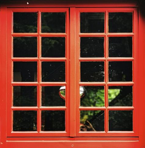 Rotes, zweiflügeliges Fenster mit je zehn quadratischen Scheiben pro Fensterflügel, die durch schmale, ebenfalls rote Rahmen voneinander getrennt sind. Hinter dem Fenster hängt mittig ein chromsilber glänzendes Herz am Rahmen, gehalten vom Fenstergriff. Die Scheiben spiegeln die grüne Umgebung und zeigen teilweise Bäume und Blätter.